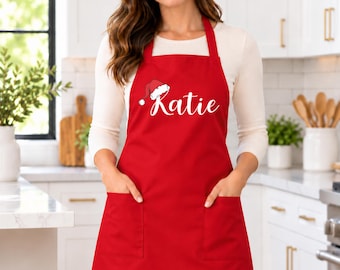 May include: A bright red apron featuring two front pockets and a white graphic of a Santa hat above the name Katie in white cursive script. The apron has a neck strap and waist ties, providing full coverage for the torso and upper legs.
