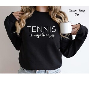 A woman wearing a black sweatshirt with the text "TENNIS is my therapy" printed on it, holding a white mug in her right hand.