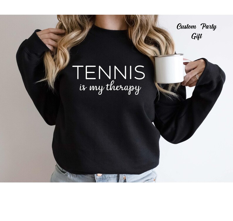 A woman wearing a black sweatshirt with the text "TENNIS is my therapy" printed on it, holding a white mug in her right hand.