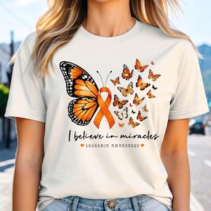 May include: A cream-colored t-shirt featuring a large orange monarch butterfly wing attached to an orange awareness ribbon. Smaller orange butterflies surround the design. The text reads I believe in miracles and LEUKEMIA AWARENESS with two orange hearts.