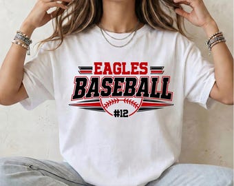 May include: A white short-sleeve t-shirt featuring a graphic print with the text EAGLES BASEBALL in red and black lettering. The design includes a stylized baseball illustration and the number 12 centered below the text.