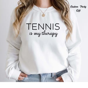 A person wearing a white sweatshirt with the text "TENNIS is my therapy" printed on it.
