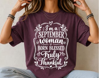 May include: A dark maroon short-sleeve t-shirt featuring white graphic text that reads I'm a SEPTEMBER woman BORN BLESSED Truly Thankful, surrounded by a decorative frame of leaves and small hearts printed on the front of the garment.