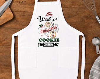 May include: A white apron featuring two front pockets and a graphic design that reads The West's CHRISTMAS COOKIE COMPANY in black, red, and green text, accompanied by illustrations of a star-shaped cookie, a snowman cookie, and holly berries.