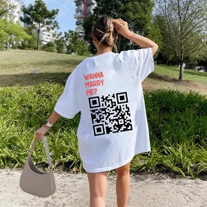 Peut inclure: T-shirt blanc surdimensionné avec le texte "Wanna Marry Me?" et un code QR noir et blanc imprimé au dos.