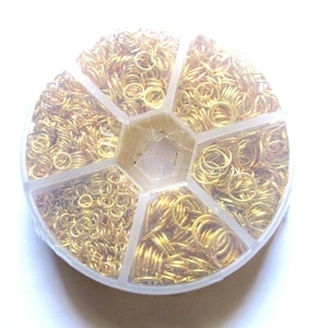 May include: A clear plastic container filled with gold-colored jump rings. The container is divided into six sections, each holding a different size or quantity of the rings. The jump rings are small, circular, and appear to be made of metal.