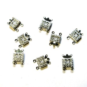 May include: A collection of silver-colored jewelry clasps. Each clasp is square with a decorative openwork design and small loops for attaching to jewelry. The clasps have a crown-like detail on top.