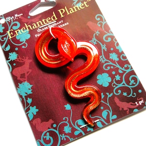 May include: A red glass pendant in the shape of a snake, with gold accents, on a maroon card with teal floral and animal designs. The card reads "Enchanted Planet" and "Glass Pendant".