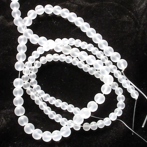 May include: Several strands of translucent, frosted white beads are arranged against a dark background. The beads vary in size, creating a textured appearance. The strands are strung together, suggesting they are ready for jewelry making or crafting.