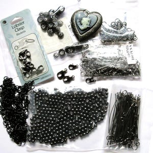 May include: An assortment of jewelry-making supplies, including lobster clasps, chains, beads, and decorative charms. The items are in various shades of black and silver, with some packaged in clear plastic bags. A heart-shaped cameo is also visible.