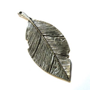 May include: A silver-colored metal feather pendant with detailed veining. The feather has a hole at the top for hanging and a slightly distressed edge. The pendant is set against a white background.