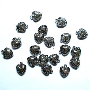 May include: A collection of small, silver-colored heart-shaped charms. Each charm features a heart design in the center, surrounded by an ornate border. They have a small loop at the top for attaching to jewelry or other crafts. The charms are scattered on a white surface.