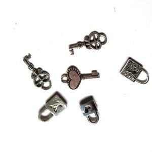 May include: An assortment of small, silver-toned charms, including keys and padlocks, are scattered on a white surface. The keys have ornate designs, and one features a heart-shaped head. The padlocks have simple, square shapes.