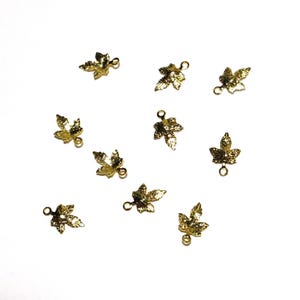 May include: A collection of ten small, gold-colored leaf charms, each with a loop for attaching to jewelry or other crafts. The charms are scattered on a white background, showcasing their intricate leaf vein details.