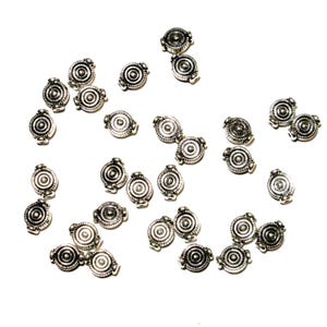 May include: A collection of small, silver-colored metal beads with a circular design. Each bead has a central circle surrounded by concentric rings and decorative edges. The beads are scattered on a white background.