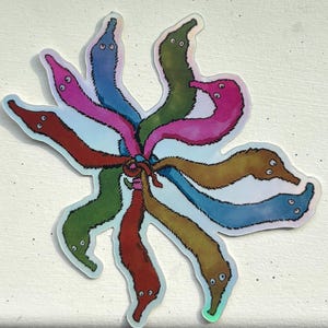 May include: A colorful sticker featuring a cluster of cartoon worms in various colors, including pink, green, blue, and brown. The worms are arranged in a circular pattern, with a holographic border. The sticker is on a white surface.
