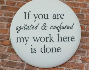 May include: A white, circular button with black text that reads, "If you are agitated & confused my work here is done." The button is set against a brick wall background.