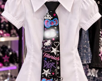 Może przedstawiać: Biała koszula z krótkim rękawem i kołnierzykiem oraz czarny krawat. Krawat ozdobiony jest grafikami Hello Kitty, srebrną koroną, zawieszkami w kształcie gwiazdek, agrafką oraz napisem Tauriah Dior w różowo-fioletowych literach z dodatkiem cyrkonii.