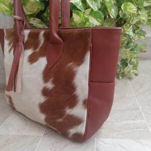 May include: A brown and white cowhide tote bag with a brown leather handle and a brown leather pocket on the side.