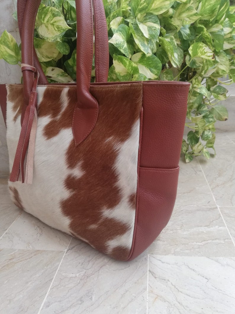 May include: A brown and white cowhide tote bag with a brown leather handle and a brown leather pocket on the side.