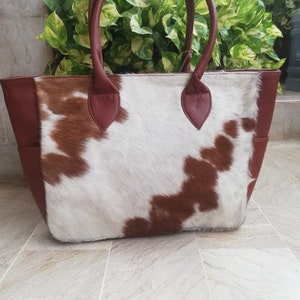 May include: A brown and white cowhide tote bag with brown leather handles and trim. The bag has a large main compartment and a smaller pocket on the front.
