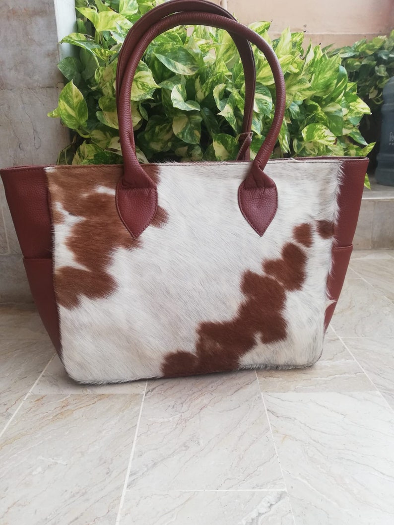 May include: A brown and white cowhide tote bag with brown leather handles and trim. The bag has a large main compartment and a smaller pocket on the front.