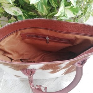 May include: A brown leather tote bag with a white and brown cowhide pattern. The bag has a zipper closure and a brown leather handle.