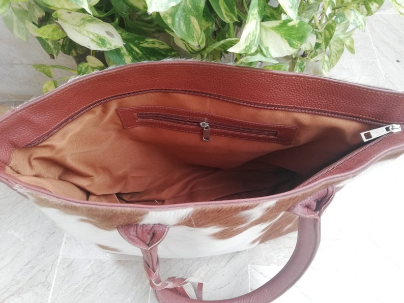 May include: A brown leather tote bag with a white and brown cowhide pattern. The bag has a zipper closure and a brown leather handle.