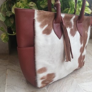 May include: A brown and white cowhide tote bag with a brown leather trim and a tassel. The bag has a top zipper closure and two top handles.
