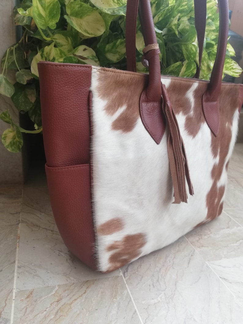 May include: A brown and white cowhide tote bag with a brown leather trim and a tassel. The bag has a top zipper closure and two top handles.