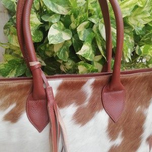 May include: A brown leather tote bag with a cowhide pattern. The bag has a brown leather handle and a brown leather tassel.