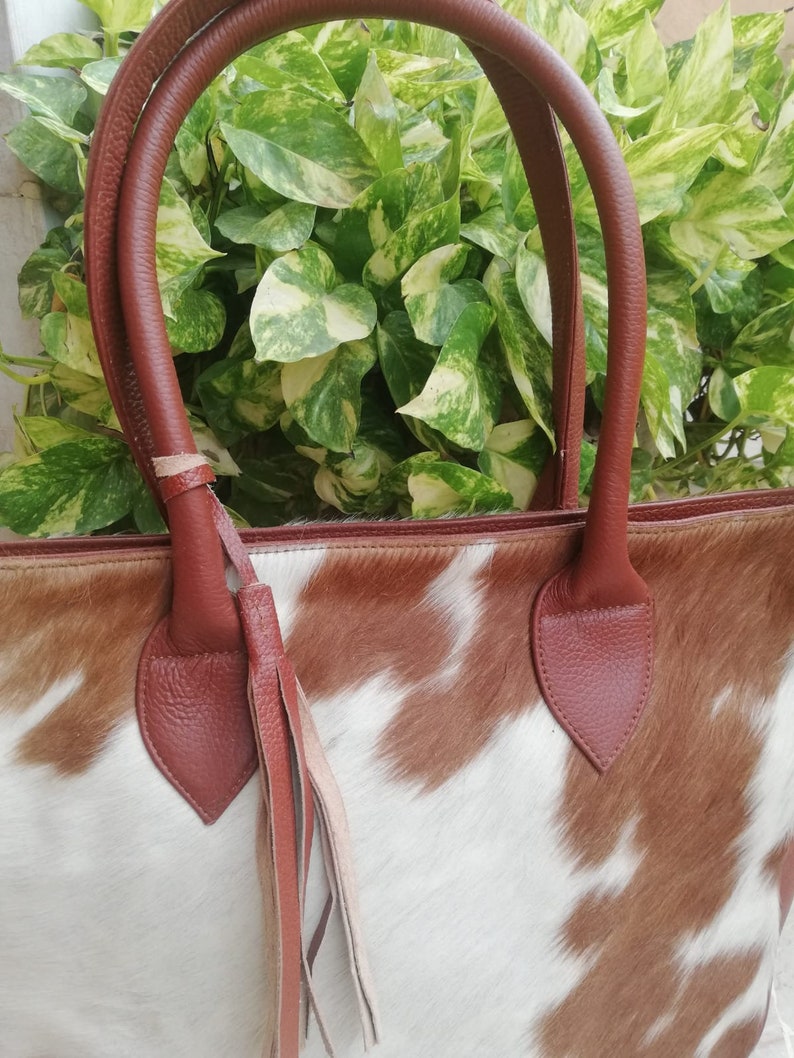 May include: A brown leather tote bag with a cowhide pattern. The bag has a brown leather handle and a brown leather tassel.