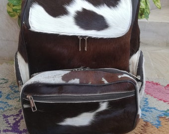 Puede incluir: Una mochila de piel de vaca marrón y blanca con una bolsa más pequeña a juego. La mochila tiene cierre de cremallera y correas ajustables.