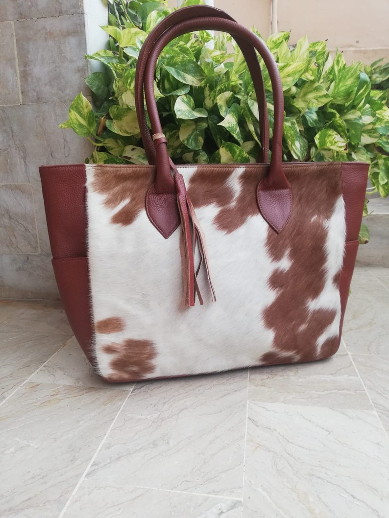 May include: A brown and white cowhide tote bag with brown leather handles and a tassel. The bag has a brown leather trim around the top and bottom.