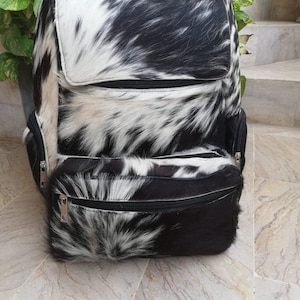 Puede incluir: Una mochila de piel de vaca negra y blanca con un bolsillo frontal con cremallera y dos bolsillos laterales.