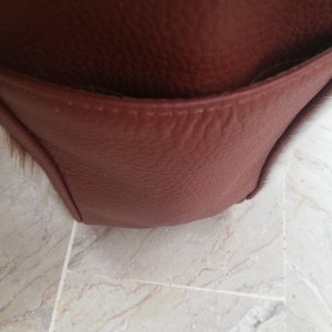 May include: Close-up of a brown leather bag with a flap pocket. The bag has a textured surface and a stitched seam.