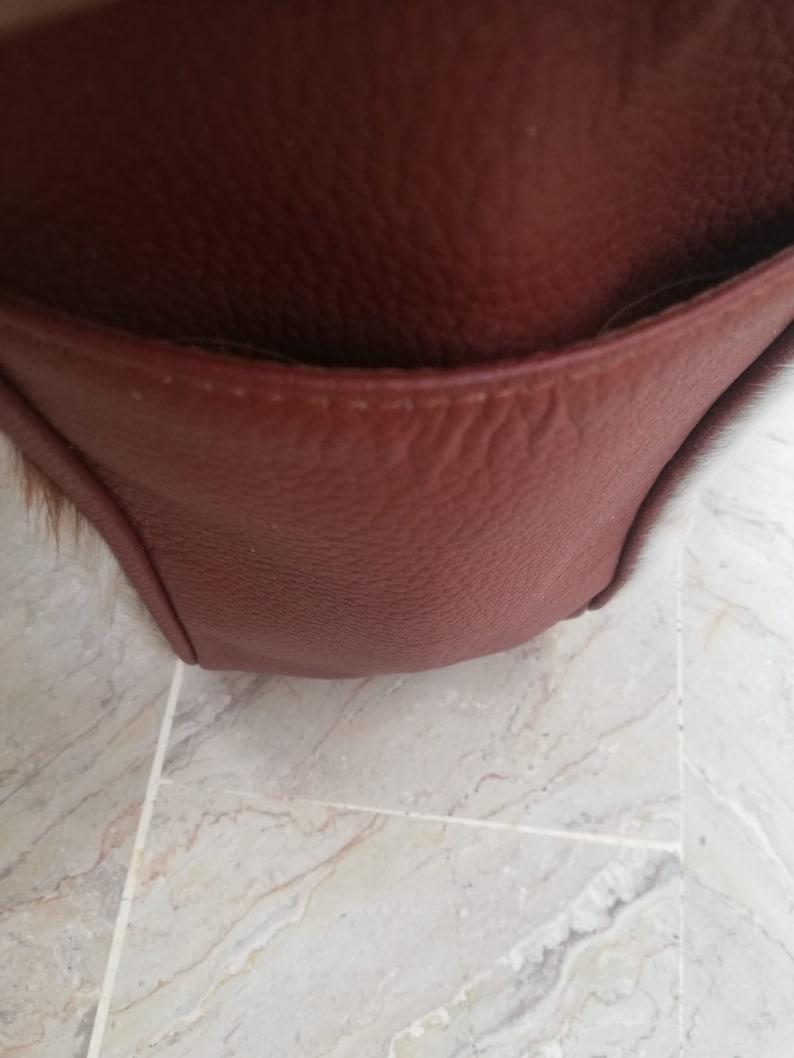 May include: Close-up of a brown leather bag with a flap pocket. The bag has a textured surface and a stitched seam.