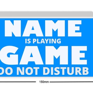 May include: A blue sign with white text that reads "NAME IS PLAYING GAME DO NOT DISTURB". The sign is 150mm wide and 80mm tall.