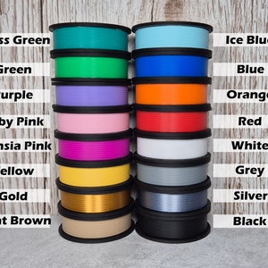 May include: A collection of 16 spools of filament for 3D printing, arranged in two rows of eight. The spools are labeled with their colors, including grass green, green, purple, baby pink, fuchsia pink, yellow, gold, light brown, ice blue, blue, orange, red, white, grey, silver, and black.