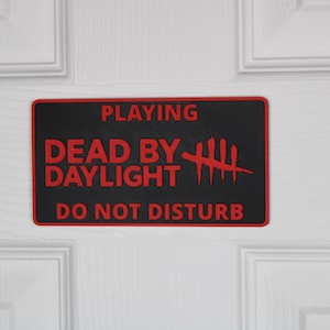 Può includere: Un cartello rettangolare nero con bordo e testo rossi. Il cartello recita "PLAYING DEAD BY DAYLIGHT DO NOT DISTURB". Il cartello è montato su una porta bianca.