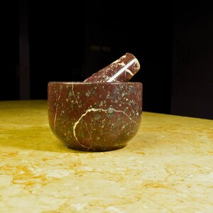 May include: A red and green marble mortar and pestle set. The mortar is a rounded bowl shape, and the pestle is cylindrical. The set is made of a stone material with a marbled pattern. The mortar and pestle are kitchen tools.
