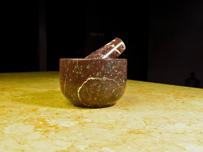 May include: A red and green marble mortar and pestle set. The mortar is a rounded bowl shape, and the pestle is cylindrical. The set is made of a stone material with a marbled pattern. The mortar and pestle are kitchen tools.