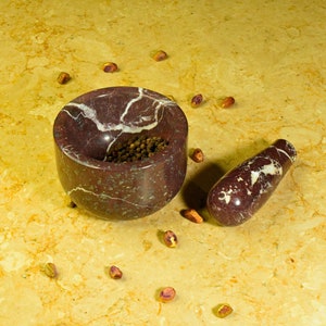 May include: A maroon marble mortar and pestle set with white veining. The mortar is filled with black peppercorns. Scattered around the set are shelled pistachios. The set is on a yellow and brown marble surface.