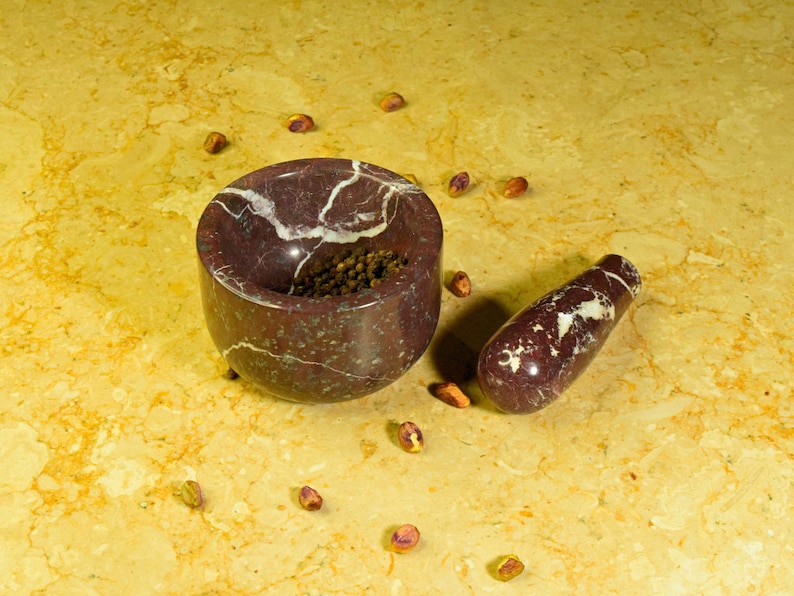 May include: A maroon marble mortar and pestle set with white veining. The mortar is filled with black peppercorns. Scattered around the set are shelled pistachios. The set is on a yellow and brown marble surface.