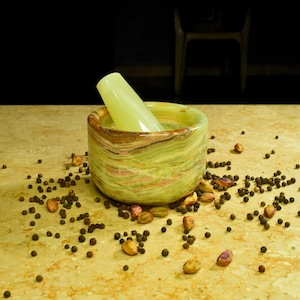 May include: A green and brown marble mortar and pestle set with a light green pestle. Black peppercorns and pistachios are scattered around the mortar on a light brown surface. The mortar is cylindrical.