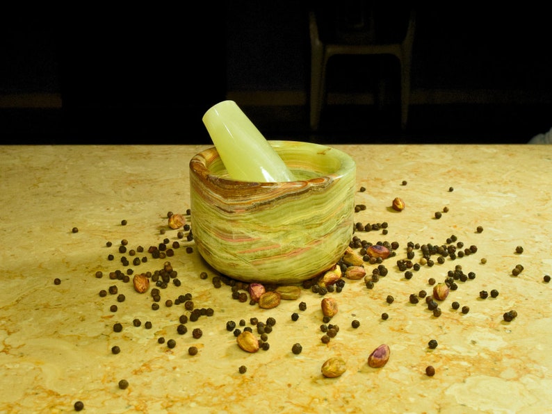 May include: A green and brown marble mortar and pestle set with a light green pestle. Black peppercorns and pistachios are scattered around the mortar on a light brown surface. The mortar is cylindrical.