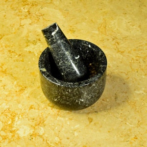 May include: A black granite mortar and pestle set. The mortar is a round bowl with a matching pestle resting inside. The set is made of dark gray stone with white veining. The background is a light yellow and brown marble surface.