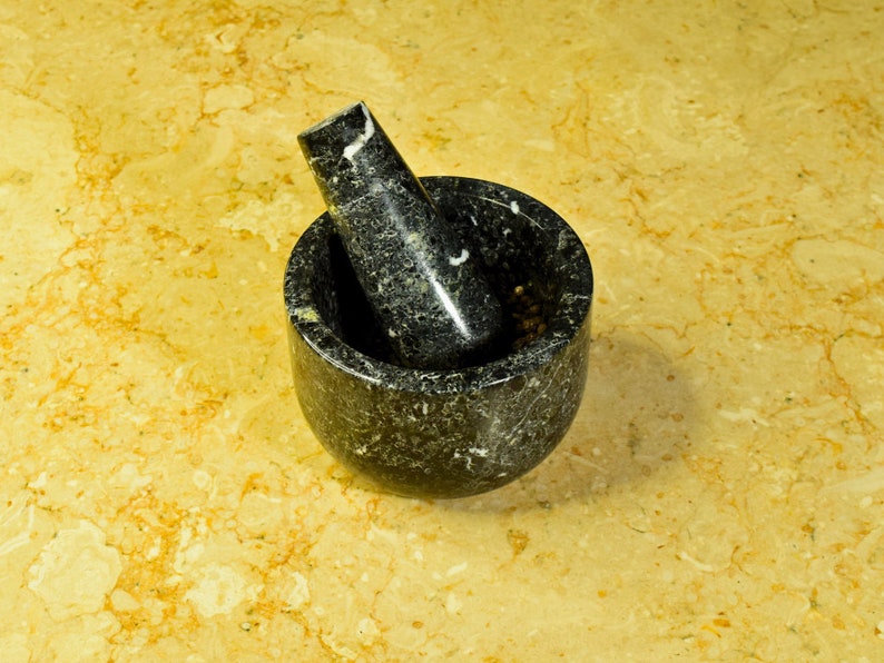 May include: A black granite mortar and pestle set. The mortar is a round bowl with a matching pestle resting inside. The set is made of dark gray stone with white veining. The background is a light yellow and brown marble surface.