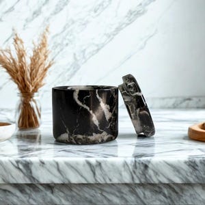 May include: A black and white marble container with a matching lid, set on a marble countertop. The cylindrical container is open, revealing its interior. A small glass vase with dried plants and a wooden bowl are also visible.