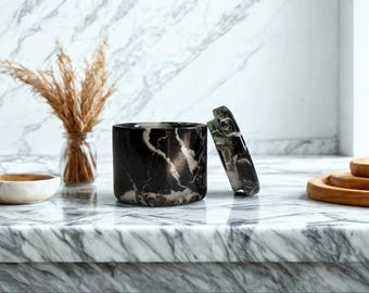 May include: A black and white marble container with a matching lid, set on a marble countertop. The cylindrical container is open, revealing its interior. A small glass vase with dried plants and a wooden bowl are also visible.